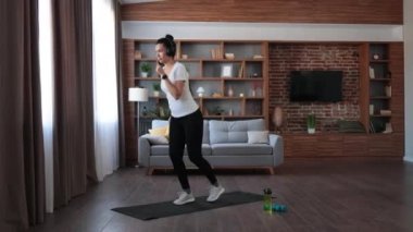 Koyu saçlı, aktif, çok ırklı bir kadın oturma odasında eğitim görürken kablosuz kulaklıkla müzik dinliyor. Spor elbiseli genç bayan spor yapmak için modern aletleri kullanıyor..