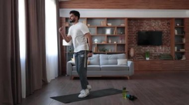 Kısa elbiseli yakışıklı Arap adam oturma odasında aktif fitness egzersizi yapıyor. Genç sakallı adam vücut şekline uygun kasları eğitiyor. Sağlıklı yaşam tarzları konsepti.