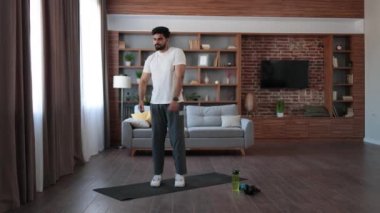 Aktif giyimli güçlü Hintli adam evde yoga minderinin üzerinde dururken kollarını esnetiyor. Yakışıklı sakallı erkek sporcu sabah vücut bakımının tadını çıkarıyor..