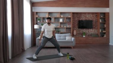 Spor kıyafetleri giymiş sakallı, yoga minderinde zıplayan, evdeki antrenman sırasında elleri başının üstüne sıkışmış Arap bir adam. Uyum, denge, ruh ve vücut bakımı için yoga pratiği.