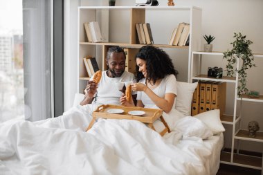 Afrika kökenli Amerikalı erkek ve kadın, güneşli bir sabahta yatakta uzanırken hilal böreği yiyip sade kahve içiyorlar. Neşeli evli çift günlük bakım yoluyla gerçek aşk mesajları gönderiyor..