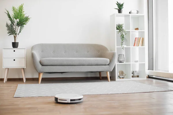 Advanced robotic vacuum cleaner hoovering airy living room in cozy apartment during sunny day. Efficient household appliance keeping hard floor and soft carpet tidy via remote control or phone app.