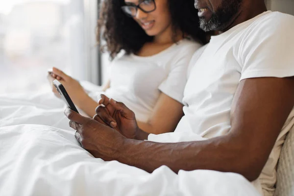 stock image Close up of fit african american male holding modern smartphone while getting rest with his wife in bed. Bearded adult man checking email messages via mobile while staying at home.