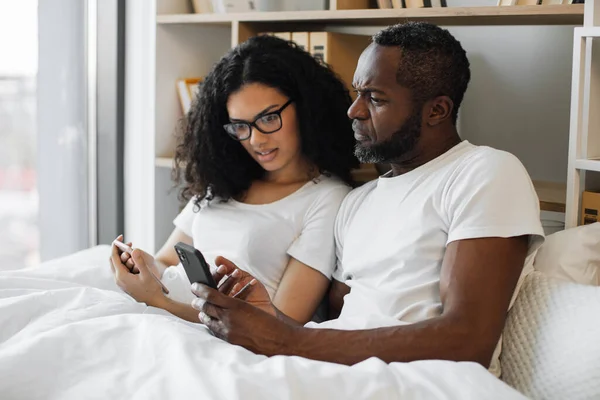 stock image Serene multiracial family of two using mobile phones while lazing in soft bed during daytime at home. Confident adult man and bespectacled woman reading online article paying attention to details.