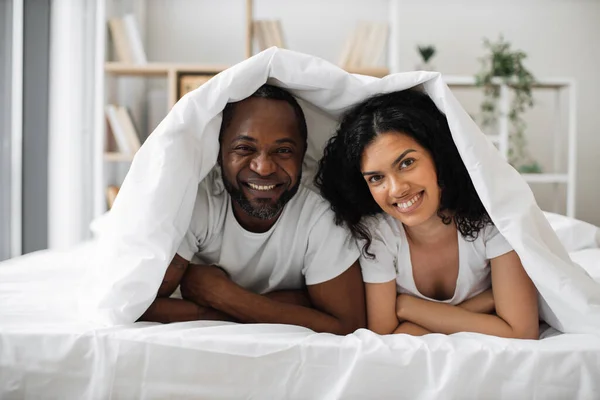 Stock image Interracial family man and woman leaning on elbows while lying under soft blanket on bed in cozy room. Happy spouses living full life by sharing intimacy and building long-lasting relationships.