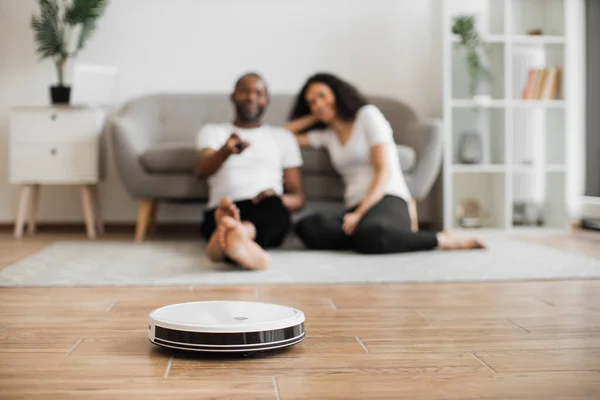 stock image Blurred background of cheerful multicultural man and woman sitting on floor and watching how wireless automatic robot cleaning their modern house. Household robots concept.