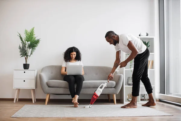 Stock image Side view of african man cleaning living room with cordless vacuum while focused woman working on laptop on couch. Caring husband doing households while wife freelancing at home.