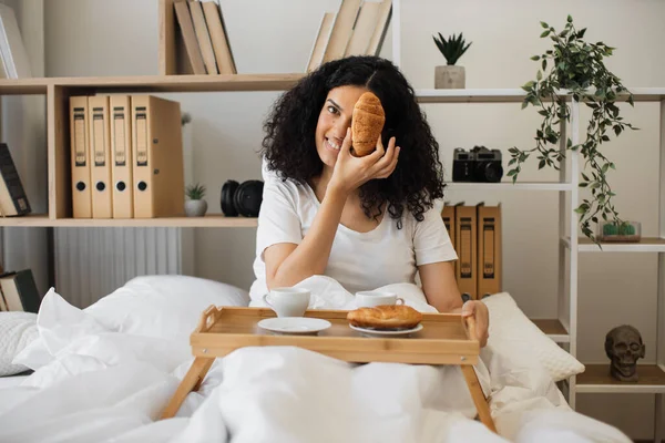 快乐的非洲裔美国女性躺在工作室公寓的床上 一只眼睛藏在月牙卷的后面 快乐的年轻女人在舒适的家享用美味的早餐 — 图库照片
