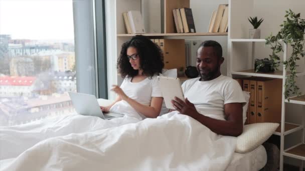 在家里躺在毯子下的多文化情侣在现代小玩艺儿上工作的前景色 通过平板电脑微笑的男人和使用笔记本电脑在网上购物的女人 — 图库视频影像
