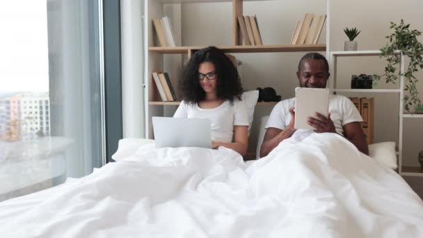 アフリカ系アメリカ人のロマンチックなパートナーは パノラマウィンドウの近くの広いベッドの上にラウンジながら 電子機器を使用しています 美しいです女性ノートパソコンとリラックスした男とタブレット楽しい時間を楽しんで自宅 — ストック動画