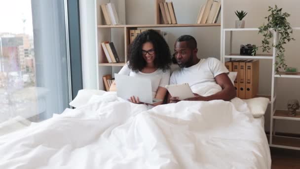 漂亮的年轻女子坐在床上 用现代笔记本电脑发短信 而她的丈夫则为她提供建议 相爱的夫妻在家中实现科技与生活的平衡 — 图库视频影像