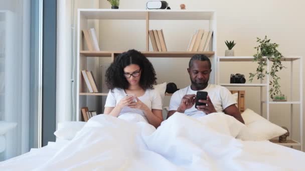 在现代公寓的卧室里 多文化的丈夫和妻子坐在舒适的羽绒被下 一边用智能手机上网一边笑 繁忙的配偶通过数字设备通过社交媒体建立联系 — 图库视频影像