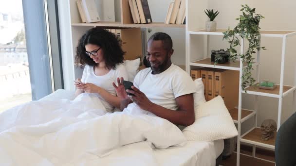 Két Tagú Nyugodt Multiracionális Család Mobiltelefonnal Miközben Otthon Puha Ágyban — Stock videók