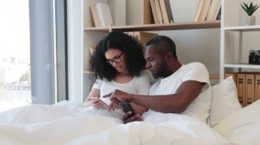 Evde gündüz vakti yumuşak yatakta tembellik ederken cep telefonu kullanan iki kişilik çok ırklı bir aile. Kendine güvenen yetişkin bir adam ve gözlüklü bir kadın online makale okuyup detaylara dikkat ediyorlar..