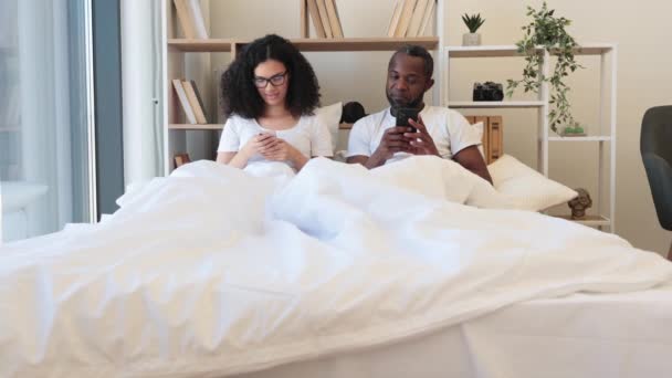 在现代公寓的卧室里 多文化的丈夫和妻子坐在舒适的羽绒被下 一边用智能手机上网一边笑 繁忙的配偶通过数字设备通过社交媒体建立联系 — 图库视频影像