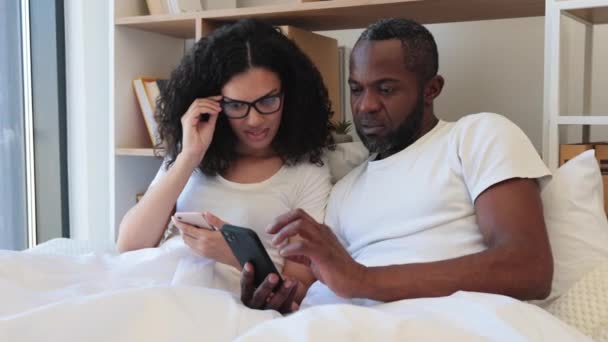 平静的多种族家庭两个使用手机 而睡在柔软的床上 白天在家里 自信的成年男人和戴眼镜的女人在网上读到关注细节的文章 — 图库视频影像