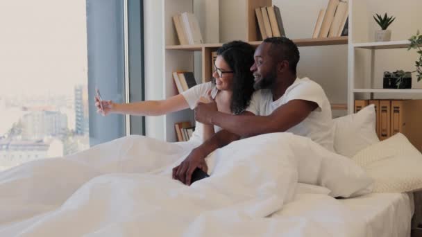 アフリカ系アメリカ人の陽気な配偶者は スタジオのアパートで休んでいる間にスマートフォンで写真を撮りました カメラで笑みを浮かべている間に親指を立てるサインを手に電話で髭を生やした男 — ストック動画