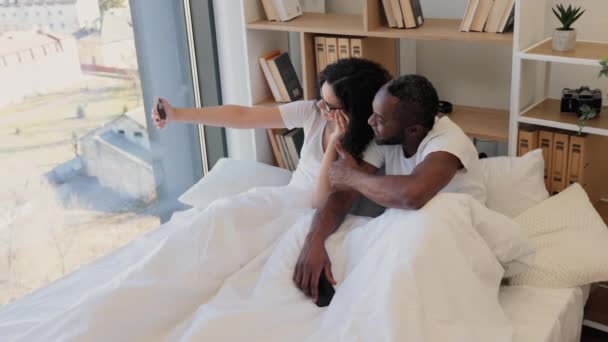 在家里躺着休息的时候 戴着眼镜笑着文化间女人 对着她和她的丈夫表现出和平的姿态 已婚夫妇在手机上拍照 以便在社交媒体上分享 — 图库视频影像