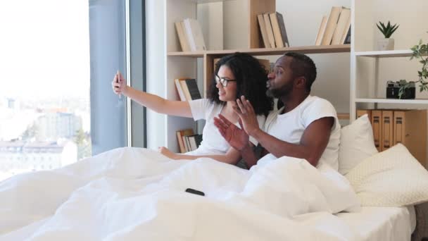 週末に寝室で楽しい時間を過ごしている間 携帯電話を使用してビデオ通話をしている楽しい陽気な人々 複数の民族の夫と妻を笑顔一緒にガジェットを使用してオンラインで話す — ストック動画