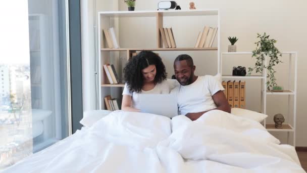 两个情绪激动的非洲裔美国家庭 一边兴奋地举起双手 一边坐在床上 带着笔记本电脑躺在毛毯上 已婚夫妇在网上成功地在家里交易 — 图库视频影像