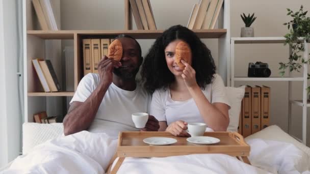 一清早 一对快乐的多民族情侣在家里享受着放在床盘上的早餐 丈夫和妻子在室内共度周末 一边笑一边用羊角面包和咖啡寻开心 — 图库视频影像