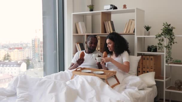 在阳光明媚的早晨 非裔美国男人和女人躺在床上 一边吃新月形的面包 一边喝着黑咖啡 快乐的已婚夫妇通过日常的照料传递着真爱的信息 — 图库视频影像