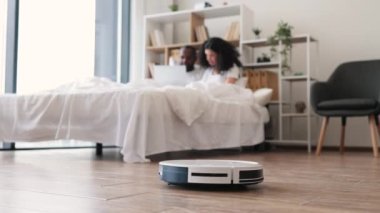 Güzel Afro-Amerikan çift rahat bir yatakta uzanıyorlar ve robotlar evlerinde yerleri süpürürken modern dizüstü bilgisayar kullanıyorlar. Modern aygıtlarla ev işleri.
