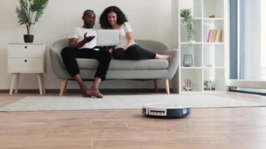 Mutlu genç çift arka planda dinlenirken otomatik robot süpürgesi ahşap zemini temizliyor. İki çok kültürlü aşık, aile yerine film izlemek için zaman harcıyor..