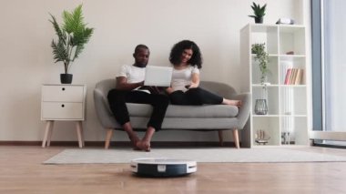 Genç ırklar arası çift rahat koltukta rahat rahat oturuyor ellerinde taşınabilir laptopları var. Kablosuz robot ahşap zemini süpürürken iki kişilik bir aile film izliyor..