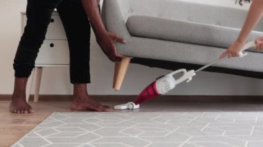 Altını süpürdüğü için sevgili karısı için gri kanepeyi kaldıran adamın ellerini kapatın. Gündelik giysiler içinde çok kültürlü bir çift boş zamanlarında modern daireyi temizliyor..
