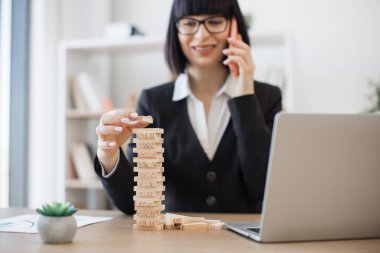 Ofis arka planında akıllı telefondan konuşurken elinde Jenga oyununun tahta bloğu olan gözlüklü beyaz bir iş kadını. İşteki kule inşaatı ile yaratıcı yönetici eğitim kontrol riski.