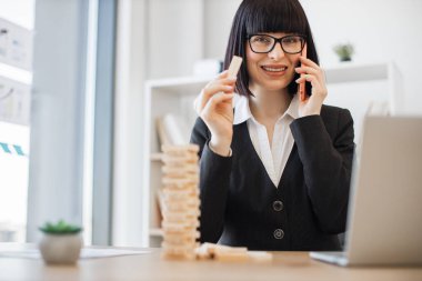Ofis arka planında akıllı telefondan konuşurken elinde Jenga oyununun tahta bloğu olan gözlüklü beyaz bir iş kadını. İşteki kule inşaatı ile yaratıcı yönetici eğitim kontrol riski.