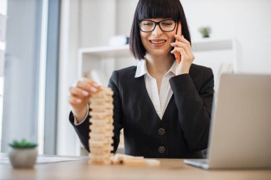 Güzel beyaz kadın resmi giyinmiş, kule inşaatı oyunuyla rahatlarken telefonla konuşuyor. Gülümseyen iş kadını ofisteki Jenga klasik oyununu kullanarak işten dolayı gerginliği azaltıyor..