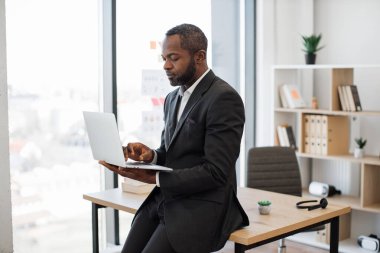 Ofis masasının kenarında dinlenirken modern dizüstü bilgisayarda çalışan deneyimli Afrikalı Amerikalı erkek işveren. Resmi giyimde odaklanmış işadamı çevrimiçi uygulama kullanarak e-postaları kontrol ediyor.