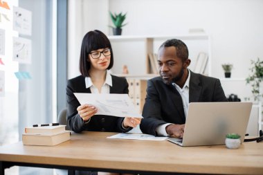 Kafkasyalı güzel iş kadını konferans masasında otururken Afrikalı Amerikalı iş arkadaşına veri görselleri gösteriyor. Tecrübeli takım arkadaşları elektronik aygıt kullanarak yıllık raporu düzenliyor.