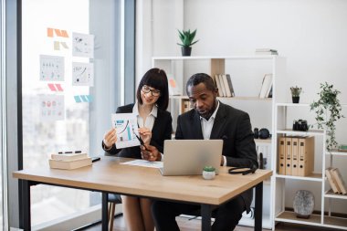 Kendine güvenen çok kültürlü bir adam dizüstü bilgisayarla iletişim kurarken beyaz bir kadın da grafikleri yükseltiyor. Verimli iş ortakları video görüşmesi sırasında projeyi tartışıyor.