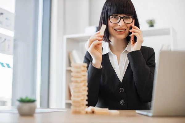 Young caucasian businesswoman in glasses holding wooden block of jenga game while speaking on smartphone on office background. Creative manager training control risk by building tower at work.