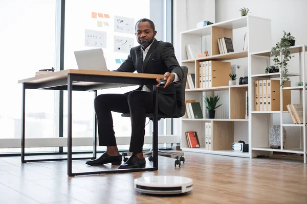 Ofis masasında otururken robot elektrik süpürgesini uzaktan kumandayla çalıştıran çok ırklı bir çalışan. Profesyonel girişimci günlük süpürge yoluyla sağlıklı iş ortamı sağlıyor..