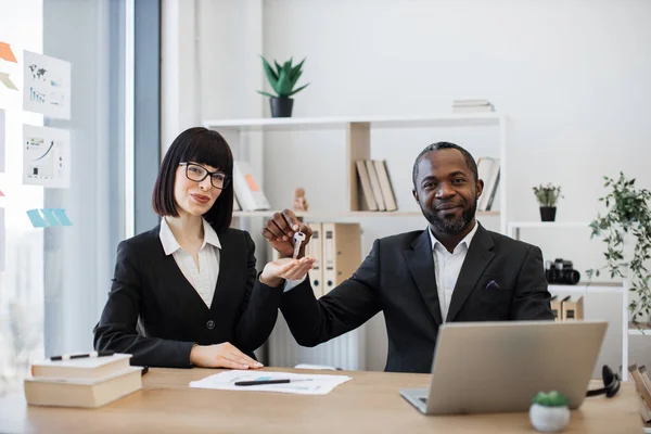 Professional managing brokers in formal clothes sitting in real estate agency with keys in hands. Cheerful caucasian businesswoman and her multiethnic male colleague celebrating successful deal.