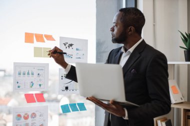 Çok ırklı ofis yöneticisi elinde bilgisayar tutarken cam tahtaya yapıştırılan çıkartmalarla ilgili kısa notlar yazıyor. Grafik ve grafiklerde finansal verilerin stil analizi.