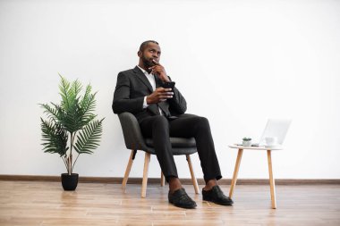 İş elbisesi giymiş Afrikalı bir adam elinde akıllı telefonuyla sandalyede oturuyor ve dikkatlice yukarı bakıyor. Düşünceli ofis çalışanı çalışma sorununu çözme fikirleri arıyor..