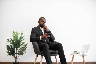 Ofiste modern akıllı telefon kullanırken dalgın bir yüz ifadesiyle Afrika kökenli Amerikalı bir adam. Takım elbiseli bir erkek işveren kahve masasının yanında dizüstü bilgisayarı ve bir bardak içkiyle oturuyor..