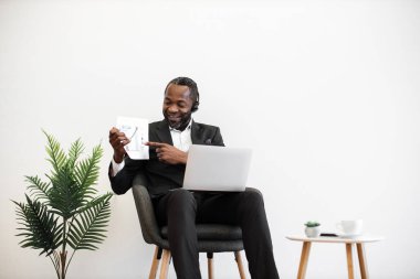 Uzman bir Afrikalı Amerikalı finansör modern laptopta online video toplantısı sırasında grafikler ve grafikler sunuyor. Resmi olarak giyinmiş, kulaklıklı bir adam ofiste oturuyor ve uzaktan görüşme yapıyor..