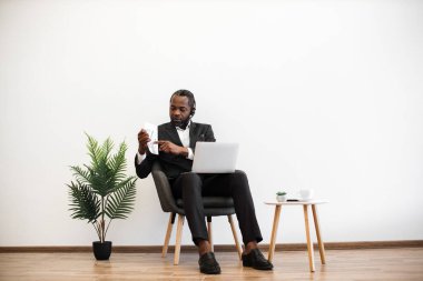 Konsantre olmuş takım elbiseli Afrikalı bir adam dizüstü bilgisayarla koltukta oturuyor ve görüntülü konuşma sırasında çizelgeleri gösteriyor. Çevrimiçi mali raporun sunumu sırasında kulaklık takan nitelikli bir ofis çalışanı.