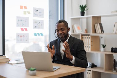 Resmi takım elbiseli Afro-Amerikan bir adam kulaklık ve kablosuz bilgisayar kullanarak ofiste video görüşmesi yapıyor. Arkaplanda çeşitli grafik ve çizelgeler olan şeffaf bir tahta. Online iş toplantısı kavramı.