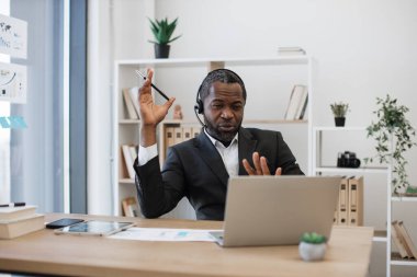 Ofis masasında oturan ve video konferansı sırasında ortaklarla konuşan kendine güvenen bir iş adamı. Afrika kökenli Amerikalı takım elbiseli bir adam uzak mesafe çalışması için kablosuz kulaklık ve laptop kullanıyor..