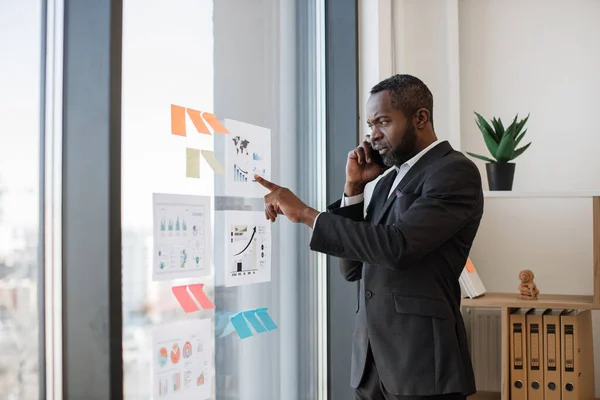 Stock image Focused multiracial businessman in smart suit receiving phone call while standing near glass panel with graphs and charts in corner office. Senior executive listening to workers speech over mobile.