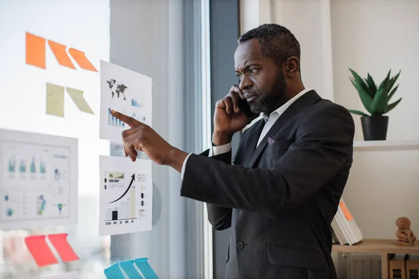 stock image Focused multiracial businessman in smart suit receiving phone call while standing near glass panel with graphs and charts in corner office. Senior executive listening to workers speech over mobile.