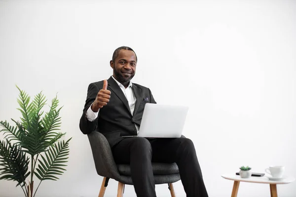 Diligent african employee in formal wear smiling sincerely at camera while typing on wireless laptop at office with modern minimalist design showing thumb up. People, work and technology concept.