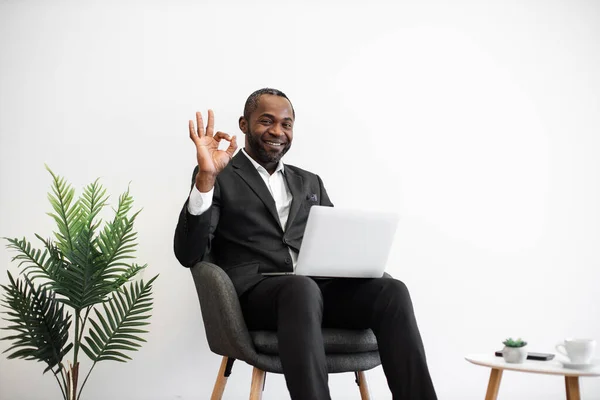 Diligent african employee in formal wear smiling sincerely at camera while typing on wireless laptop at office with modern minimalist design showing sign ok. People, work and technology concept.